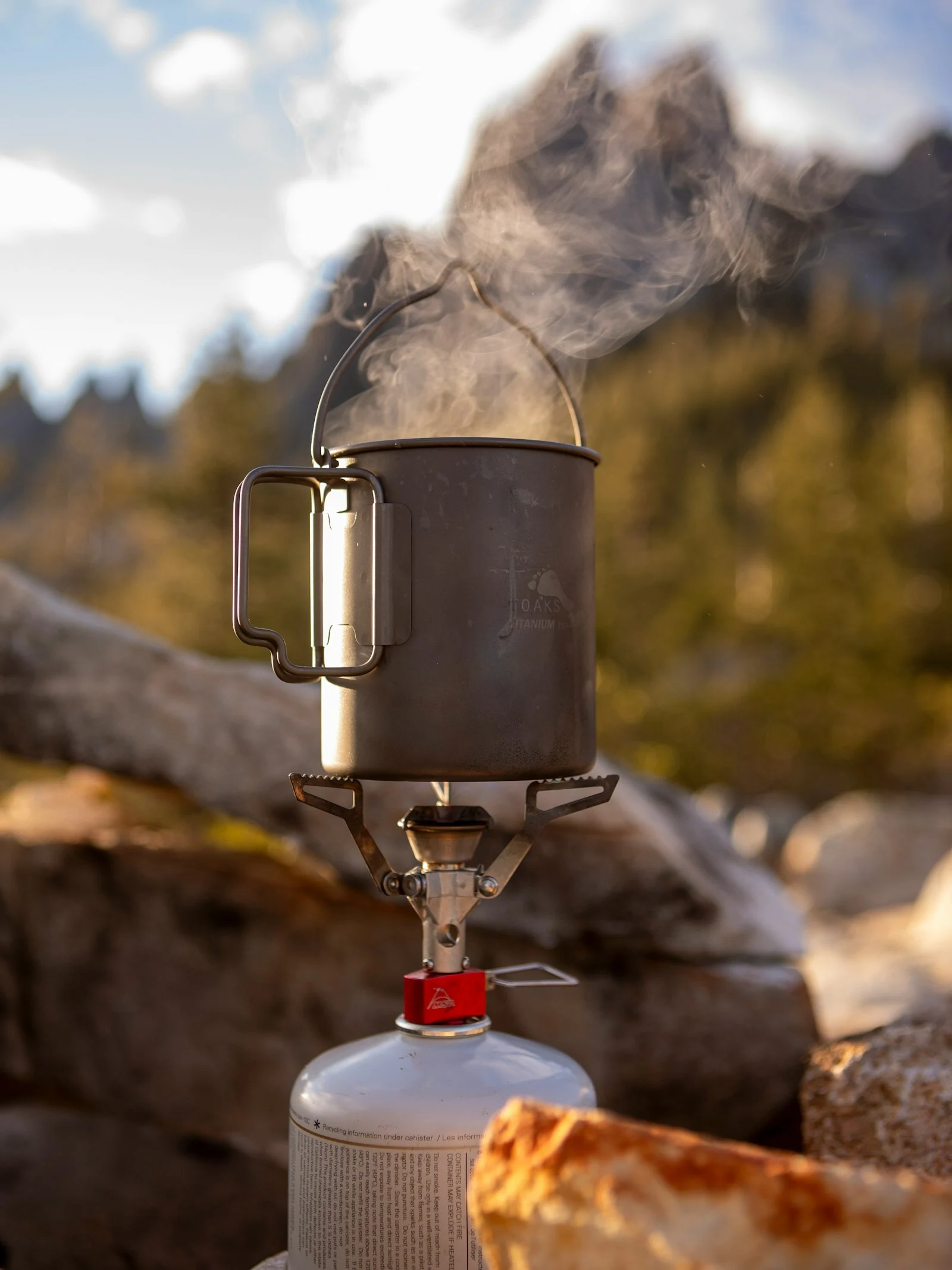 Pure titanium camping cookware pot in outdoor setting - lightweight GR1 titanium pot on campfire with mountain background Pure titanium camping cookware pot in outdoor setting - lightweight GR1 titanium pot on campfire with mountain background